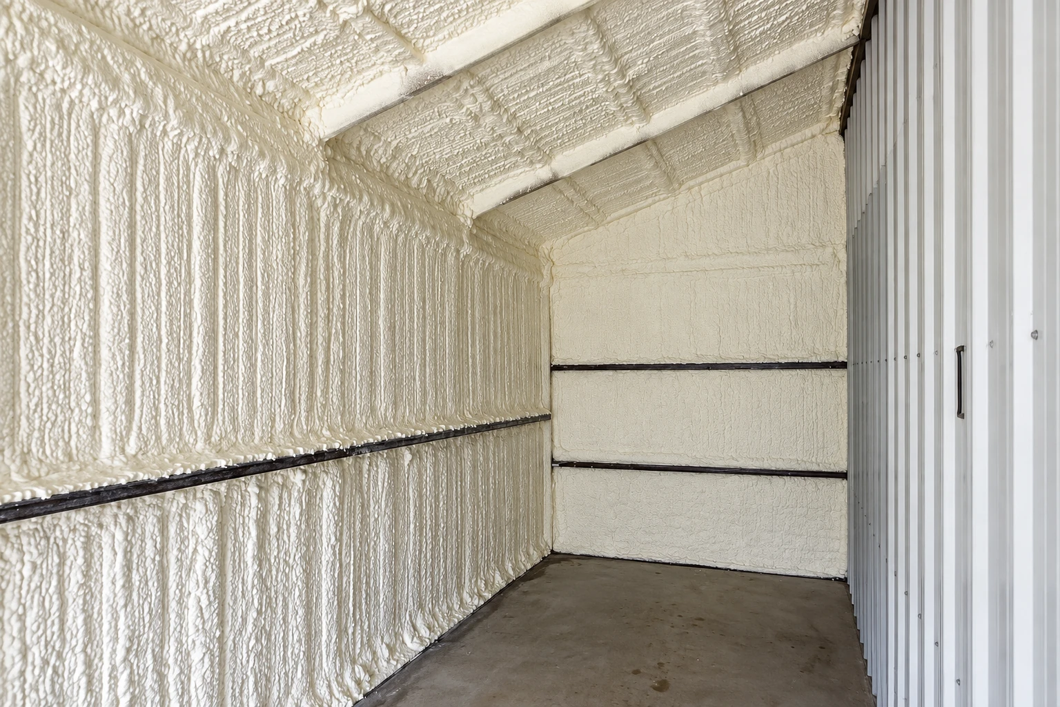 White spray foam coats the walls and ceiling of a narrow unfinished room beside a corrugated metal wall.