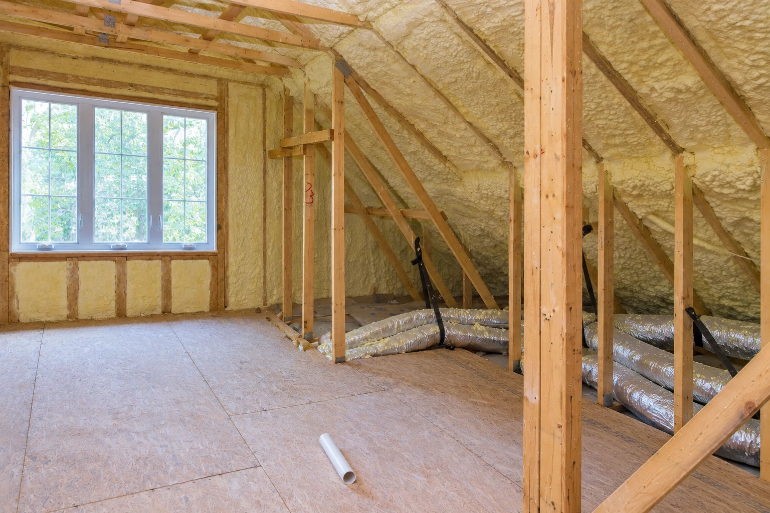 Attic rafters and sloped walls are coated in pale spray foam, above a plywood floor and flexible ducts.