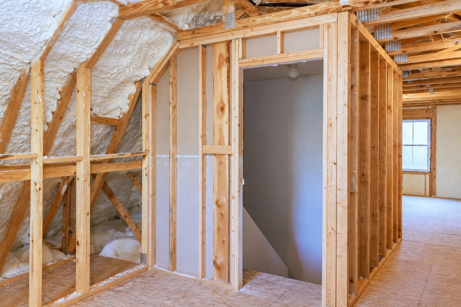 Unfinished attic with white spray foam insulation on the sloped roof framing beside exposed wooden studs and a doorway.