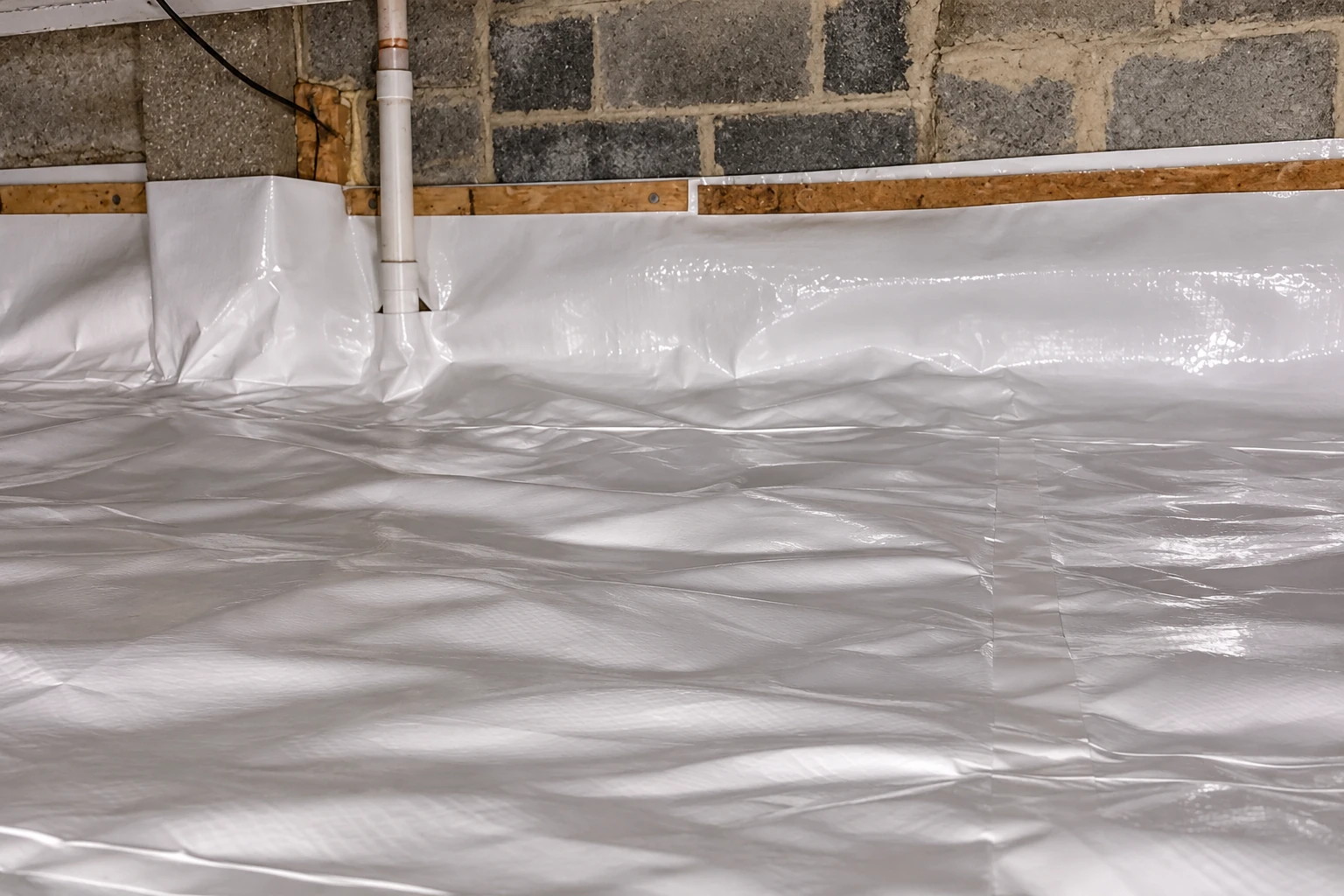 Crawl space floor covered with a white plastic vapor barrier, with the sheeting turned up along a block wall and around a pipe.