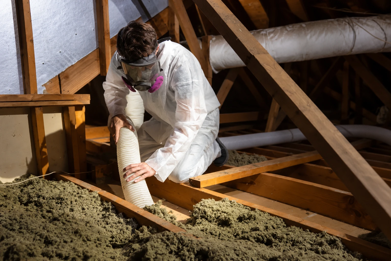 A worker wearing protective gear uses a hose to remove loose attic insulation between wooden joists.