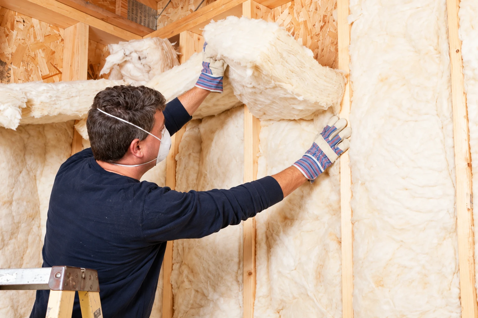 A masked worker in gloves fits fluffy fiberglass batts between exposed wooden wall studs.