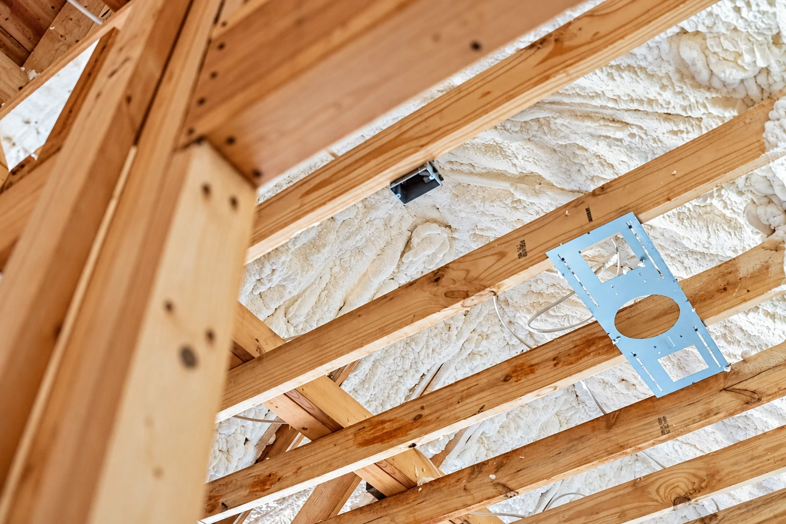 White spray foam fills the spaces between exposed wooden roof trusses, with a metal mounting plate attached.